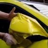 A yellow conformable cast wrapping film is being stretched and conformed over a car wing mirror. Two hands are holding the edges of the vinyl at the top and bottom and pulling it over the smooth outside face of the wing mirror.