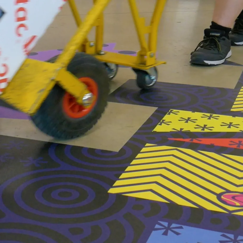 Mactac FloorGrip floor graphic laminate with an abstract colourful lineart design has been stuck to a warehouse floor and a yellow trolley with red wheels carrying two Mactac graphics boxes is seen being rolled over the floor decoration.