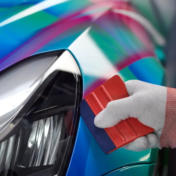 Mactac white cotton wrapping glove holding a red vinyl wrap squeegee as the self-adhesive film is pressed into the contoured edges of a car headlight.