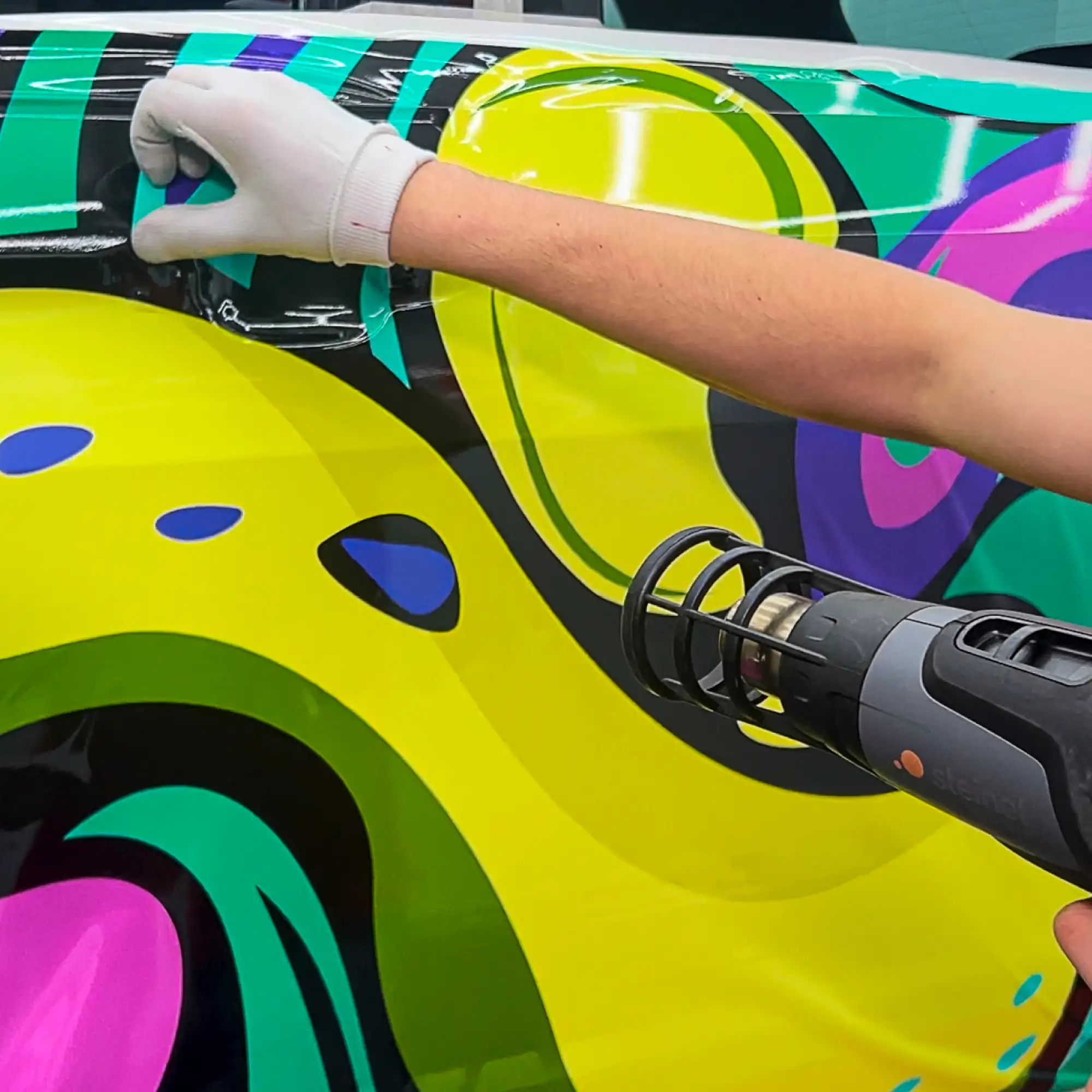 Mactac JT 10800 WG-BFG printable vehicle wrapping vinyl being installed to the side of a car with a cloe-in detail of the installer wearing a white wrapping glove and applying heat to the film with a heatgun.
