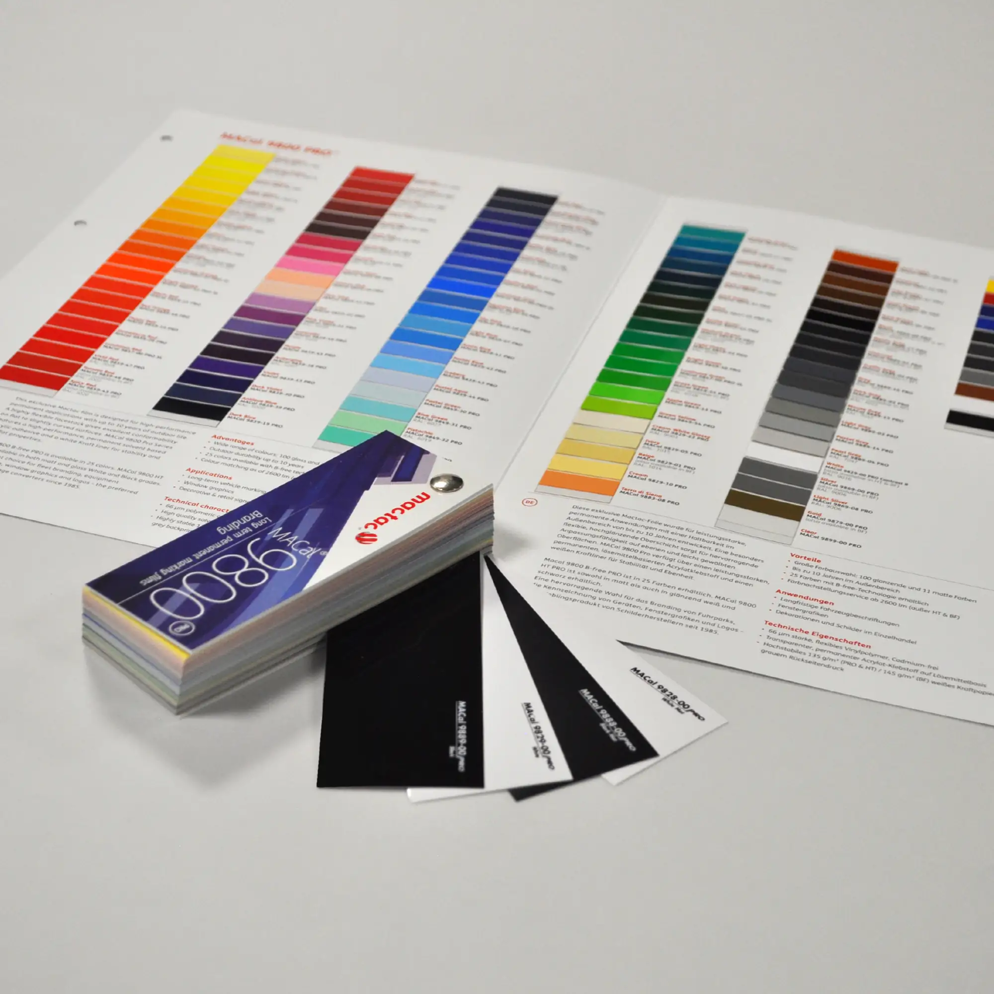 Mactac's MACal 9800 swatch, folded into a block with only the 9800 HT (High Tack adhesive) black and white, gloss and matt vinyl sample options are protruding from the swatch block. In the background is an opened colour card that shows the full range of 9800 colour self-adhesive sign vinyls.