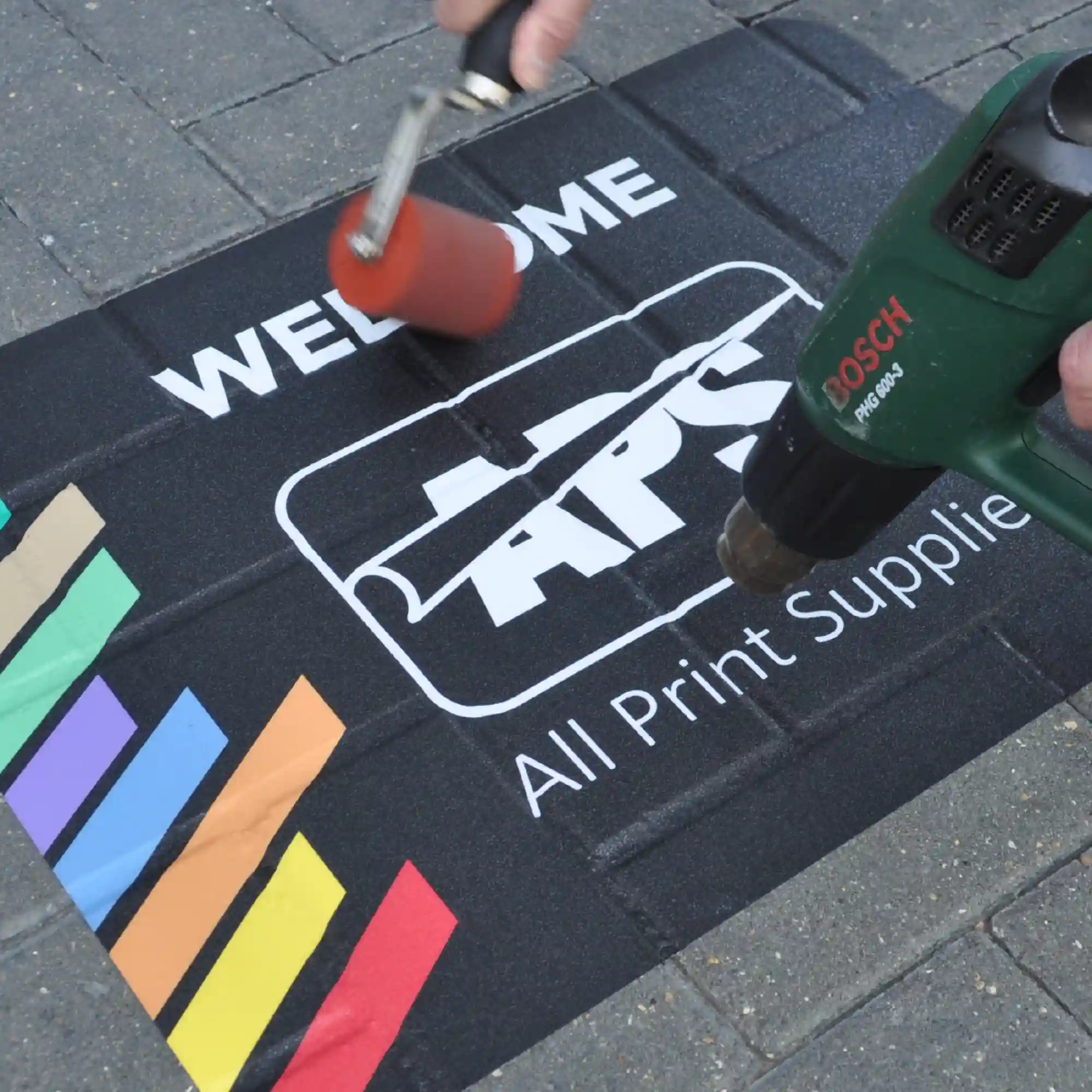 Mactac StreetRap print film laminated with StreetLam, this printed external floor entry 'welcome' graphic has been applied to block paved bricks with a vibrant printed branded APS logo image and is shown in the process of being heat and roller applied and fixed into the recess of the paving gaps.
