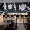Mactac WallChalker self-adhesive chalkboard vinyl applied to a wall behind a public bar seating area with printed white ink graphics of sketch style animals enjoying drinks surrounded by promotional offer text that has been written between the graphics with liquid chalk pens.