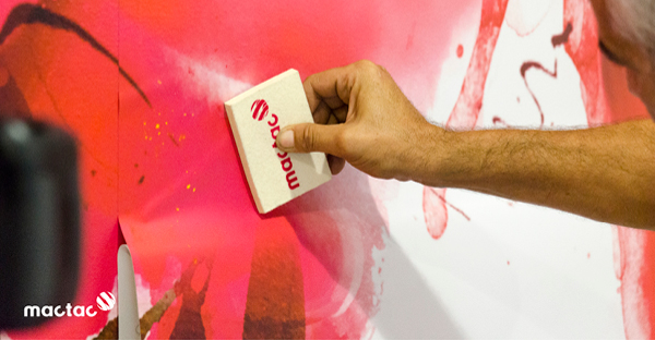 Mactac WW309 self-adhesive wall art print vinyl being applied with a Mactac branded graphics squeegee to an interior waiting room wall. Close-up of the printed red and white poppy graphic with the liner material folded down as the release paper is removed and the wallcovering is applied into position to adhere to the wall.