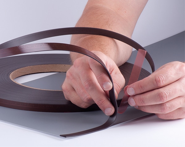 The brown coloured Magnetic Tape coil is pictured being held at the loose end of the tape as the installer releases the red PE film liner from the double-sided adhesive tape and sticks down the adhesive side of the magnetic tape to the rear of some pop-up display media laid out on a flat work bench.