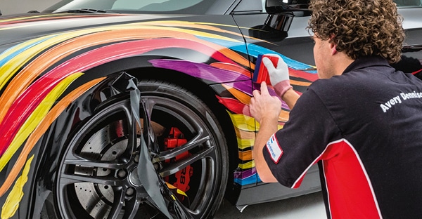 MPI 1105 EA RS comformable cast wrapping film being squeeged onto a sports car. The digitally printed colourful graphics are of a painted brushtrokes design in primary colours of cyan blue, yellow, orange, red and purple as the designs flowing lines follow and exagerate the contours of the cars elegant shape across the door, wheel arch, wing mirrors and bonnet.