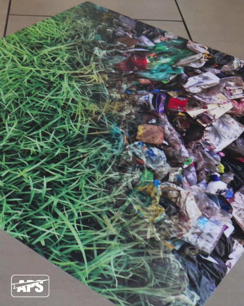 A large floor graphic applied to an indoor tiled reception floor space, printed on Mactac's JT 5425 PUV and laminated with slip resistant LUV 6400. The image shows green blades of grass merging diagonally into a second photo image of a huge pile of plastic waste and general rubbish.