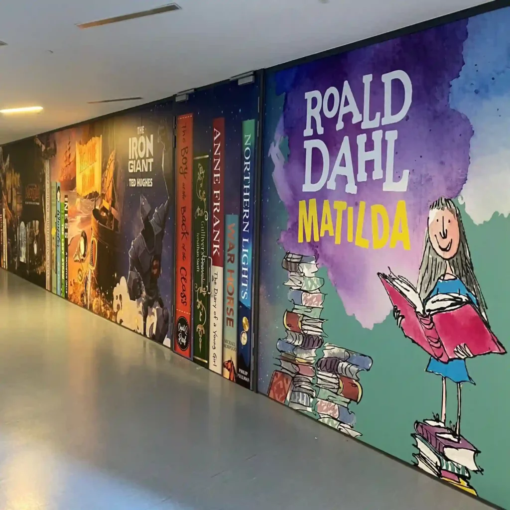 NU-COAT 'M' Series monomeric print vinyls for flat sided applications, used here adhered to a long school corridor wall with inspirational and educational imagery encouraging the enjoyment of book reading featuring a book-shelf design with large books with spines and covers on display side-by-side.
