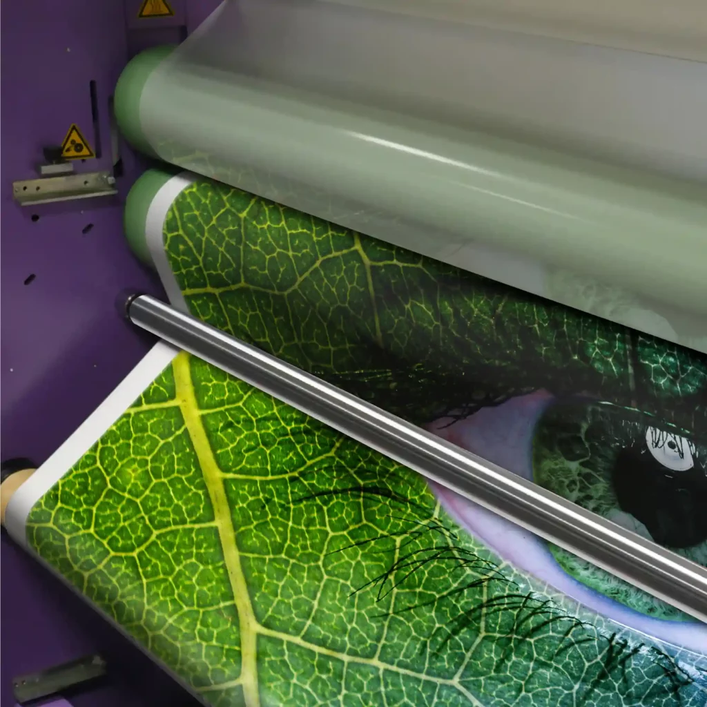 NU-COAT O21-P-P gloss clear polypropylene, PVC-free eco-print laminate with permanent self-adhesive, for flat sided graphic applications, is shown passing through mutiple rollers on a wide-format printers laminator, in the process of being overlaminated onto a green leafy eye printed image.