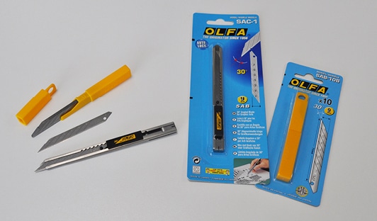 OLFA SAC-1 retractable snap-off knife in a slimline stainless steel construction with retractable blade shown with the AB-10B, 30° replacment blades in 13 segments laying next to the original OLFA packaging the knife and blades are supplied in - all the objects are fanned out in a semi-oval shape.