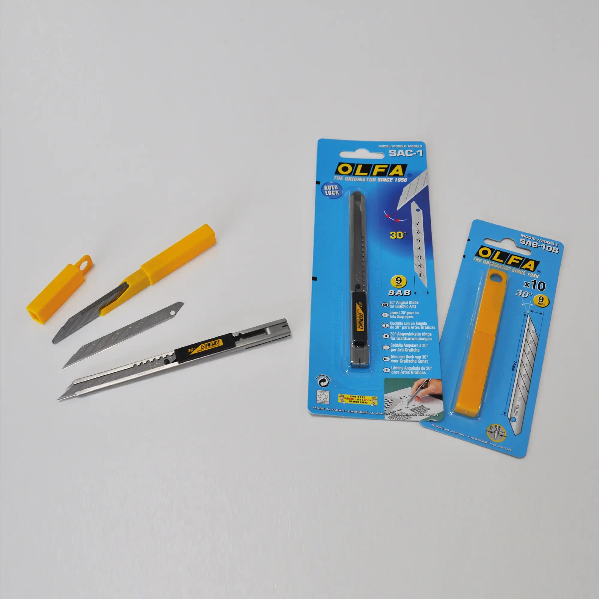 OLFA SAC-1 retractable snap-off knife in a slimline stainless steel construction with retractable blade shown with the AB-10B, 30° replacment blades in 13 segments laying next to the original OLFA packaging the knife and blades are supplied in - all the objects are fanned out in a semi-oval shape.