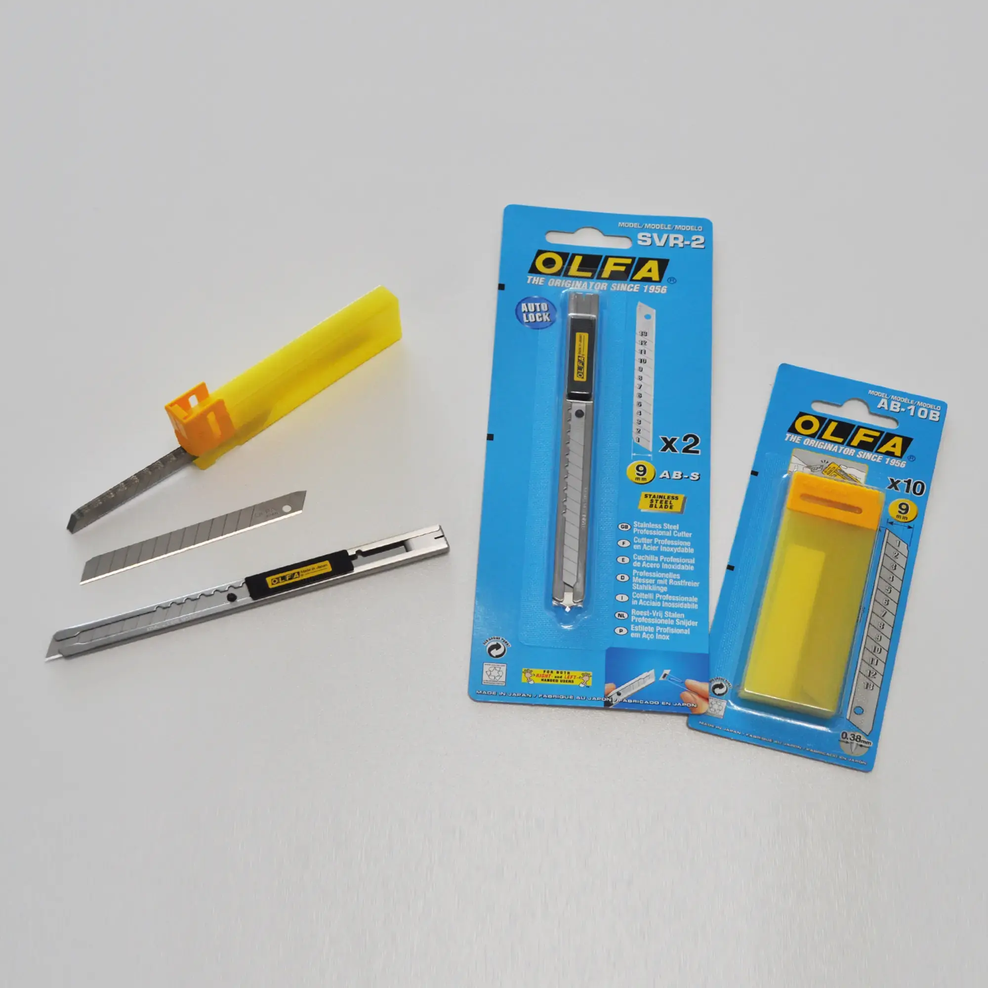 OLFA SVR-2 retractable snap-off knife in a slimline stainless steel construction with retractable blade shown with the SAB-10B, 45° replacment blades in 7 segments laying next to the original OLFA packaging the knife and blades are supplied in - all the objects are fanned out in a semi-oval shape.