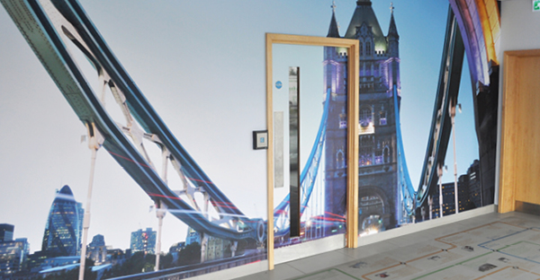 Royal Majestic 1025-11 printable wall fabric pasted to an internal plastered wall with a print of Tower Bridge in London, UK, in blue hues with red streaks as the traffic rushes by. The viewing angle is seen from pedestrian level from the centre of the bridge looking slightly upwards at one of the blockwork towers in the central distance with suspension bars coming outwards towards and away from the viewer spreading upwards to the left and right of the image.