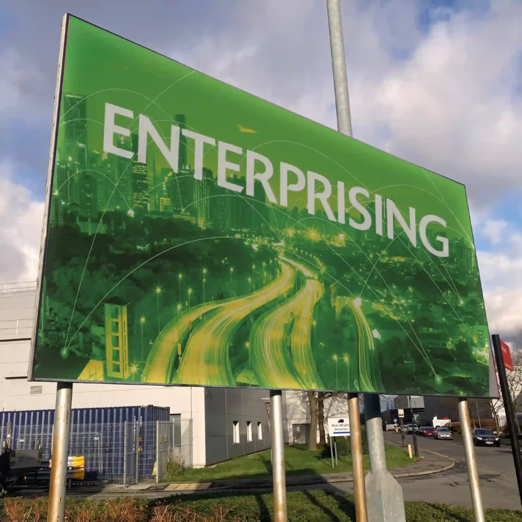 Printed billboard advertising on large-format digital premium poster paper, featuring 'enterprising' text overlaid on a green tinted photographic image of a digitally inteconnected city positioned on a busy, high mounted roadside bill-board poster sign.