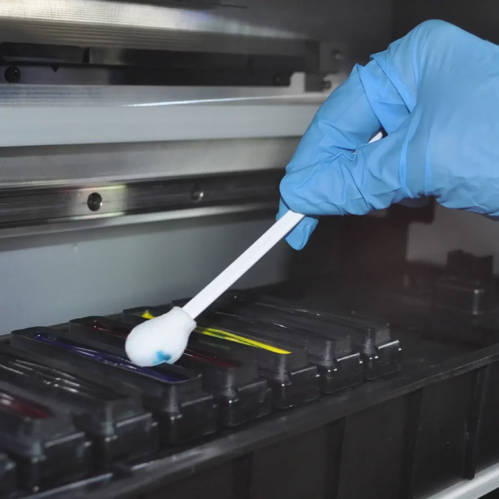 A blue gloved hand holds a foam tip cleaning swab into the printer capping station and is wiping away some excess cyan blue ink which can be seen on the foam head as it is soaking up and clearing away the ink.