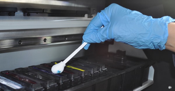 A blue gloved hand holds a foam tip cleaning swab into the printer capping station and is wiping away some excess cyan blue ink which can be seen on the foam head as it is soaking up and clearing away the ink.