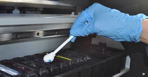 A blue gloved hand holds a foam tip cleaning swab into the printer capping station and is wiping away some excess cyan blue ink which can be seen on the foam head as it is soaking up and clearing away the ink.