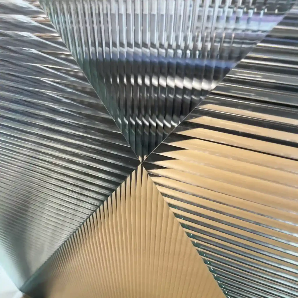 Reeded Glass window films provide decorative privacy to glass surfaces, with a subtle lens effect the clear stripes are available in two widths which are pictured here applied to a large glass partition wall where the 2 different lens sizes have been cut into symetrical triangle pieces and re-assembled to form an "X" shape with the slimmer RG6 pattern film positioned vertically and the wider RG12 lens width applied horizontally, both meeting at a point in the middle of the film. The obscurity of the films are apparent as the photo looks through the films and the view behind is obscured as it is broken up into sections where the light is bent giving a shallow ribbed effect.