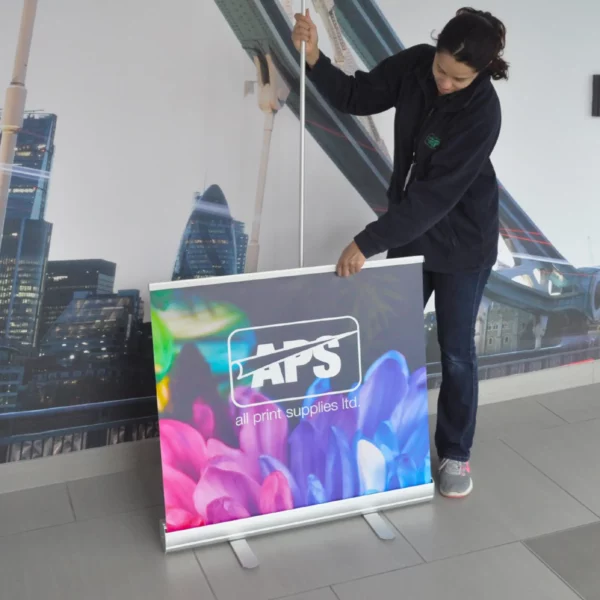 Pulling up the printed roll-up display banner graphic with it extended half-way showing the support pole behind and the cassette and 2 x extended feet at the bottom of the photo.