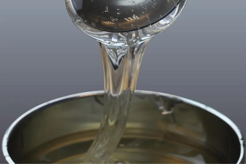 Clear viscous, reasonably runny, polyacrylate self-adhesive graphic adhesive being ladled out of a tin to show the warmed up liquid slowly glooping back downwards into the tin.