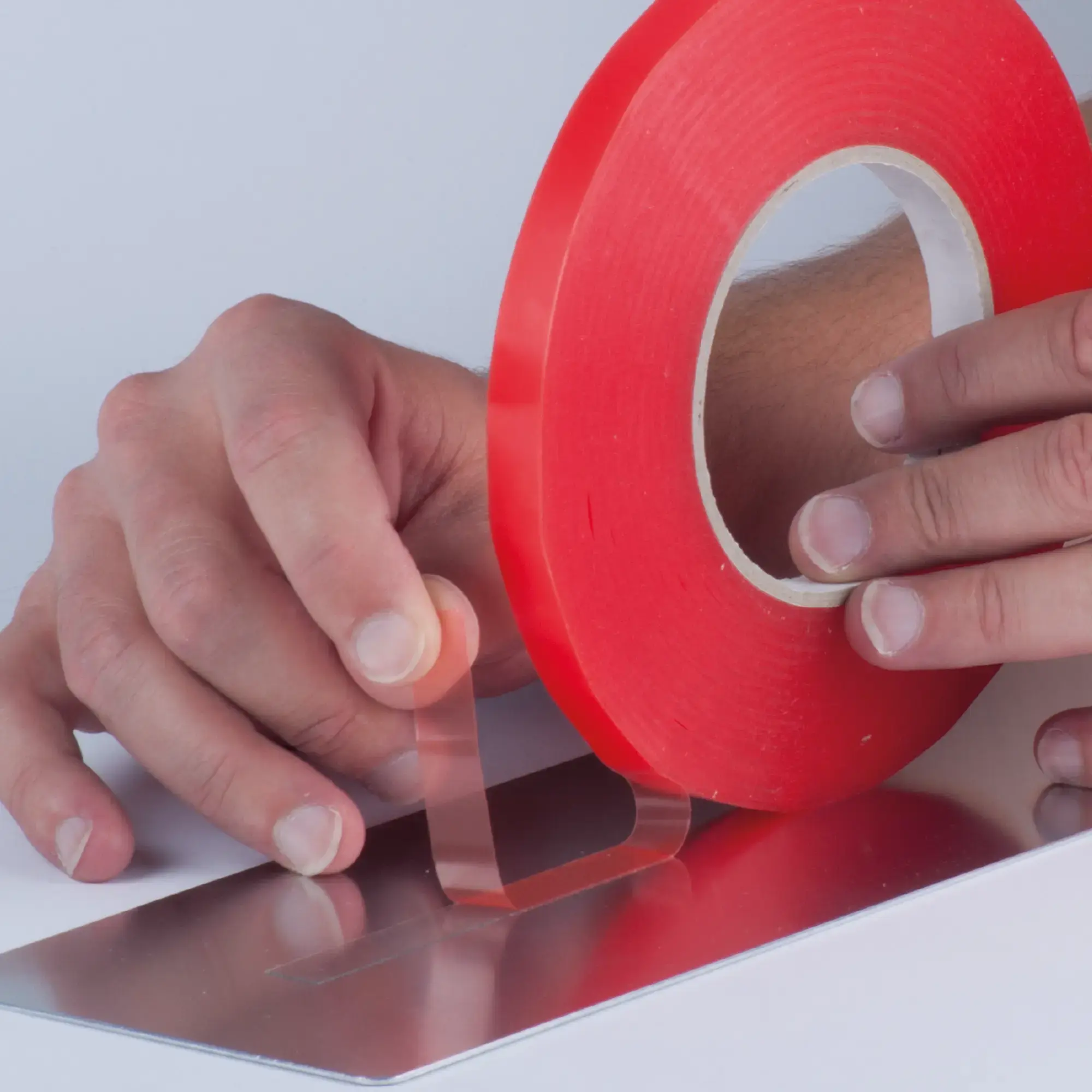 The outside (top) protective liner of DS-C-2065F clear double-sided sign tape is being peeled away to expose the adhesive on the top side as the tape on the other side (bottom) is adhered in a straight line to a Di-Bond metal sheet on a work bench.