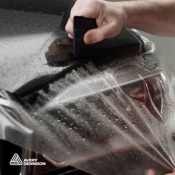 A close in view of Avery Dennison SPF being squeegeed onto the front side of a bonnet surface, with the film being pulled into position over the top of the vehicles headlight.