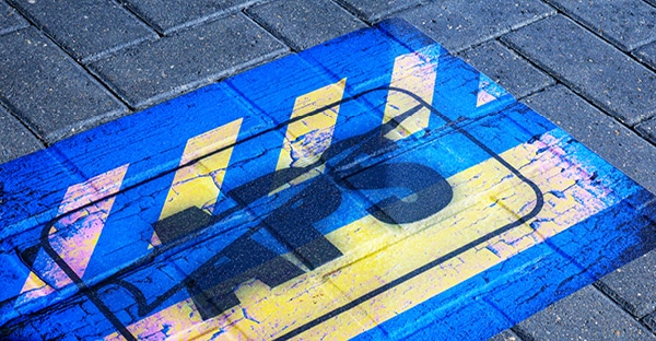 Streetrap print vinyl laminated with StreetLam for the highest rated slip resistant floor graphics has a rustic broken pavement image with stripes of blue and yellow hues and a black APS logo emblazended centrally on top of the image. The graphic is adhered in a car park area onto a rough, bitchumen surface.
