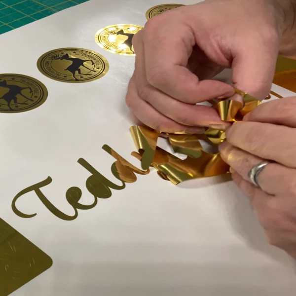 Weeding away the waste film from NU-COAT T61-P bright, two-way gold self-adhesive sign vinyl to finish some print n cut graphics of reindeer postal stamp circles and personalised text lettering graphics.
