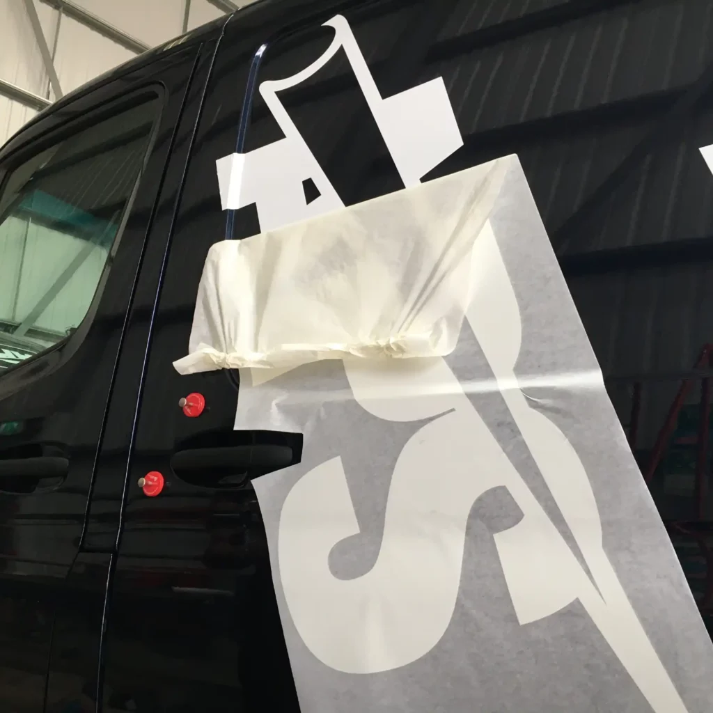 TT60 application tape is holding cut-vinyl van livery in position on the side of a van, whith the app tape half-peeled back, away from the self-adhesive sign vinyl logo graphic leaving the logo in perfect position and keeping all separate vinyl parts together during the application process.