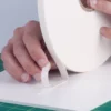 The outside (top) protective liner of the white FM-W-10P foam tape is being peeled away to expose the adhesive on the top side as the sign tape as the other side (bottom) of the tape is adhered in a straight line to a foam board sheet on a work bench.