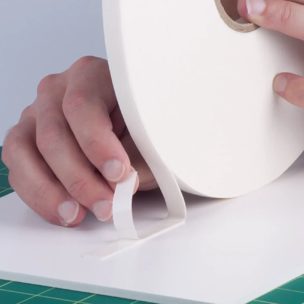 The outside (top) protective liner of the white FM-W-10P foam tape is being peeled away to expose the adhesive on the top side as the sign tape as the other side (bottom) of the tape is adhered in a straight line to a foam board sheet on a work bench.