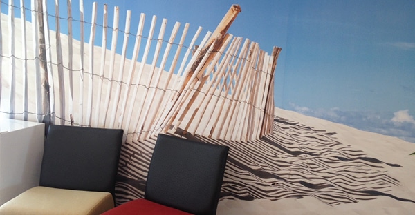 WW 200 wall covering media applied to a smooth waiting room wall with a a printed scene of sand dunes with a wooden fence casting a shadow on the ripples in the sand with a clear blue sky. You can see the context of the waiting room the wall wrap vinyl is hung in as the waiting room chairs are in front of the wall graphic.